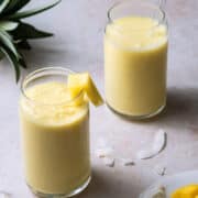 Two glasses of mango smoothie without yogurt are surrounded by pineapple.