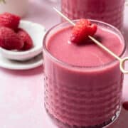 A skewered raspberry is laying on top of a glass of raspberry smoothie.
