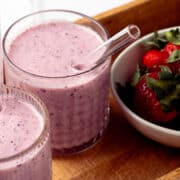 A bowl of strawberries is next to two glass with a strawberry blueberry smoothie.