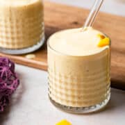 A small glass has the apple mango smoothie with a glass straw in the glass and a wooden cutting board around the back.