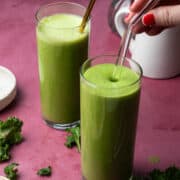 Two mango kale smoothies are on a purple background with a hand holding a straw.