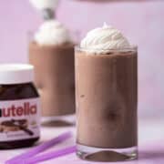 Whipped cream tops a Nutella frozen hot chocolate. A hand is piping whipped cream on top of the milkshake is the background.