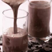 The blended chocolate smoothie is being poured into a glass with a full glass behind it.