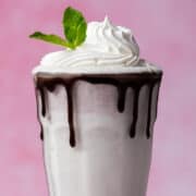 A mint chocolate chip milkshake has chocolate syrup dripping off of the glass with a mint sprig in the whipped cream.