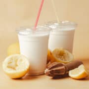 Two whipped lemonades are in plastic cups surrounded by lemon pieces and a juicer.