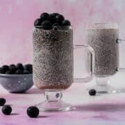 Two blueberry chia seed puddings are in mugs with handles and blueberries are around the mugs.