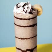 The chocolate banana milkshake is in a tall glass with a blue background on a yellow table.