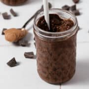 A spoon of peanut butter is behind a jar of chia seed pudding surrounded by chocolate chunks.