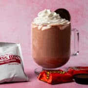 A frozen peanut butter hot chocolate is topped with whipped cream and surrounded by peanut butter cups and packets of hot chocolate.