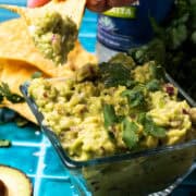 A chip is taking some of the 4 ingredient guacamole out of the square bowl.