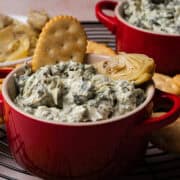 An artichoke and a cracker is on top of the cold spinach and artichoke dip in a red bowl.