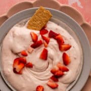 A small piece of graham cracker is in a bowl with the strawberry cheesecake dip and topped with chopped strawberries.