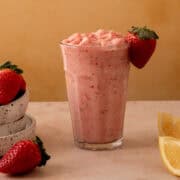 A bowl of strawberries is next to a glass full of strawberry frozen lemonade.