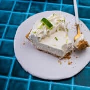 A slice of the no bake key lime pie is on a white plate on a blue background with a fork holding a piece of the pie.