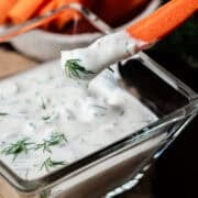 A carrot stick is being dipped into the square bowl full of the Greek yogurt ranch dressing.
