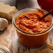 A spoon is in a jar of the roasted red pepper pesto with parmesan cheese in the background.