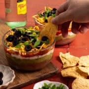A hand is scooping the seven layer dip out of the container.