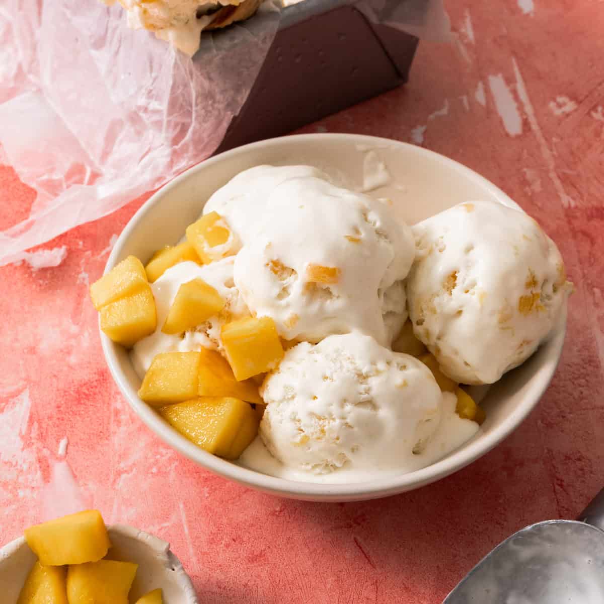 Three scoops of no churn mango ice cream are in a bowl with chunks of mangoes in the bowl.