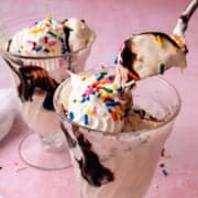 A spoon is lifting the no churn vanilla ice cream out of a glass topped with sprinkles and hot fudge.