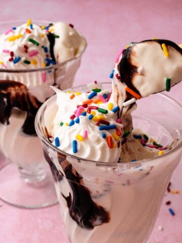 A spoon is lifting the no churn vanilla ice cream out of a glass topped with sprinkles and hot fudge.