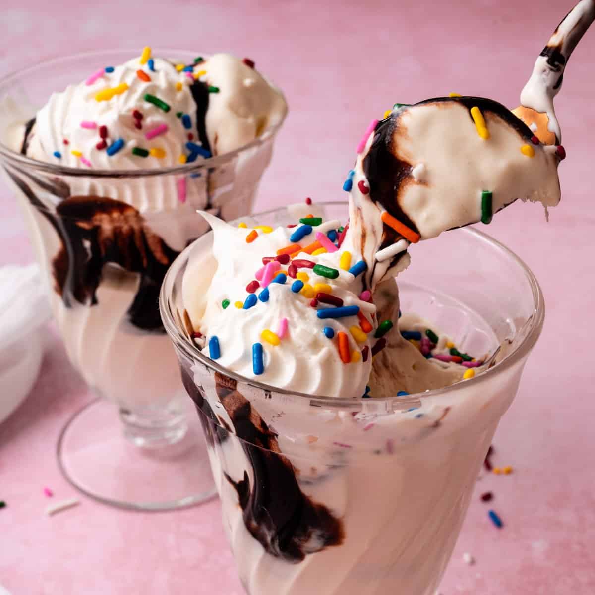 A spoon is lifting the no churn vanilla ice cream out of a glass topped with sprinkles and hot fudge.
