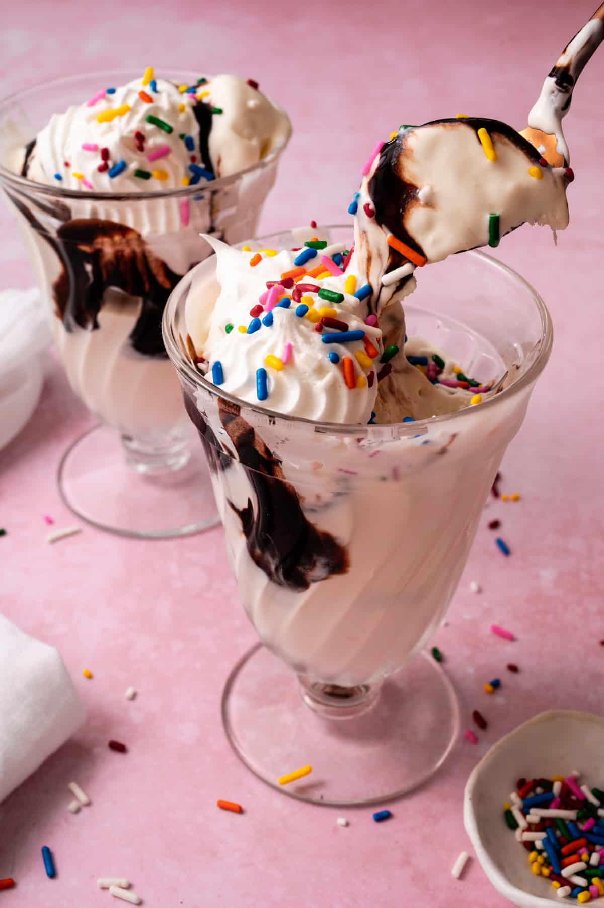 A spoon is lifting the no churn vanilla ice cream out of a glass topped with sprinkles and hot fudge.