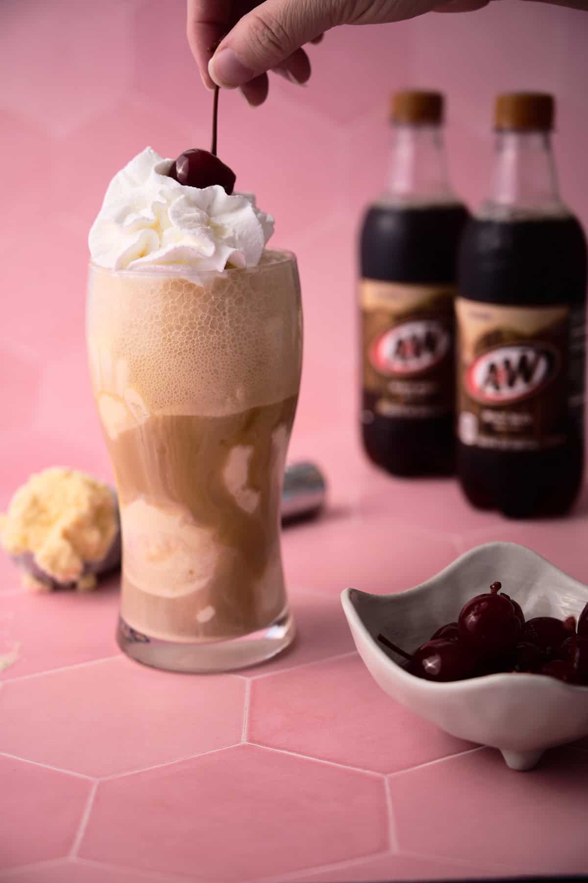 A hand is lifting the cherry off of the glass of root beer float.
