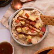 A graham cracker is added to the bowl of the apple cinnamon dip that is topped with chopped apples.