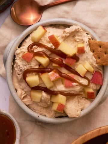 A graham cracker is added to the bowl of the apple cinnamon dip that is topped with chopped apples.