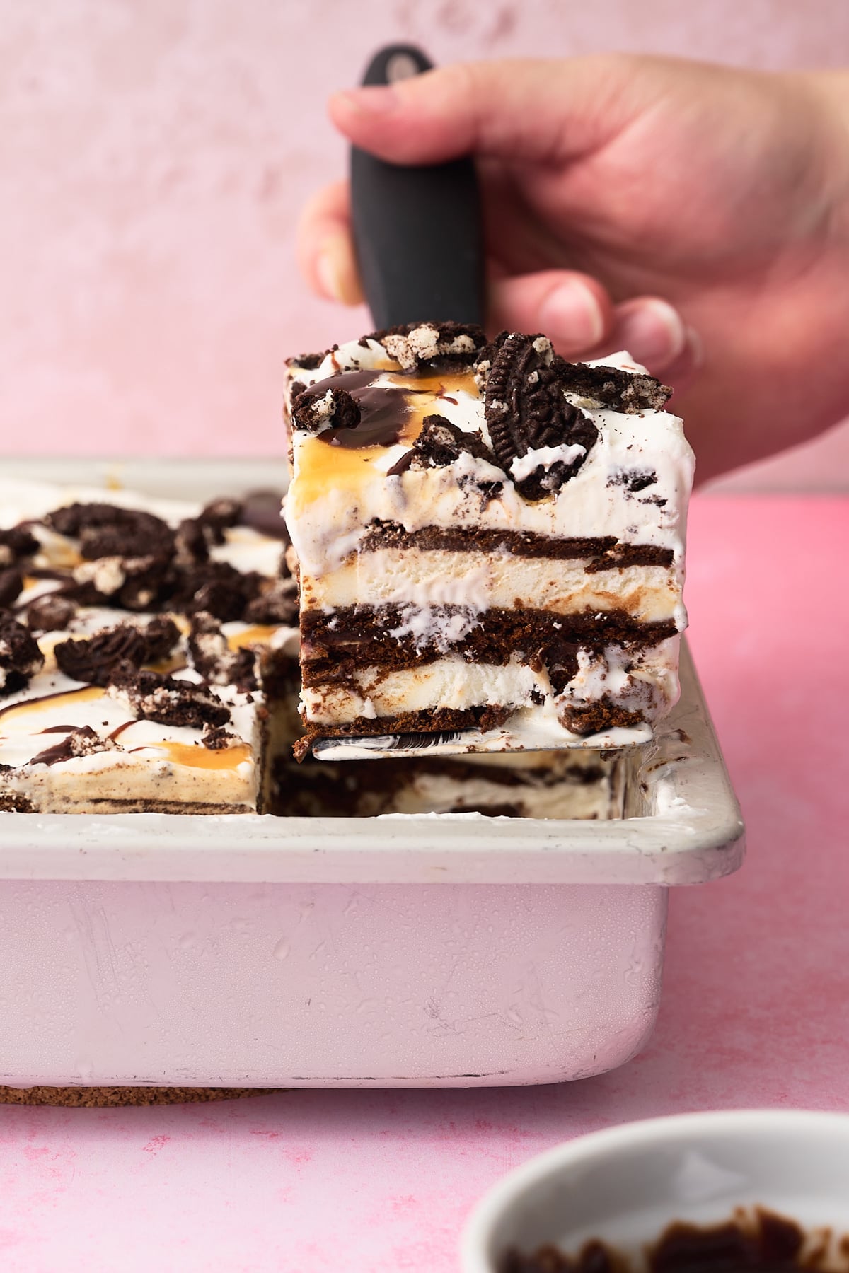 A slice of the ice cream sandwich cake is being taken out of the pan with a hand.