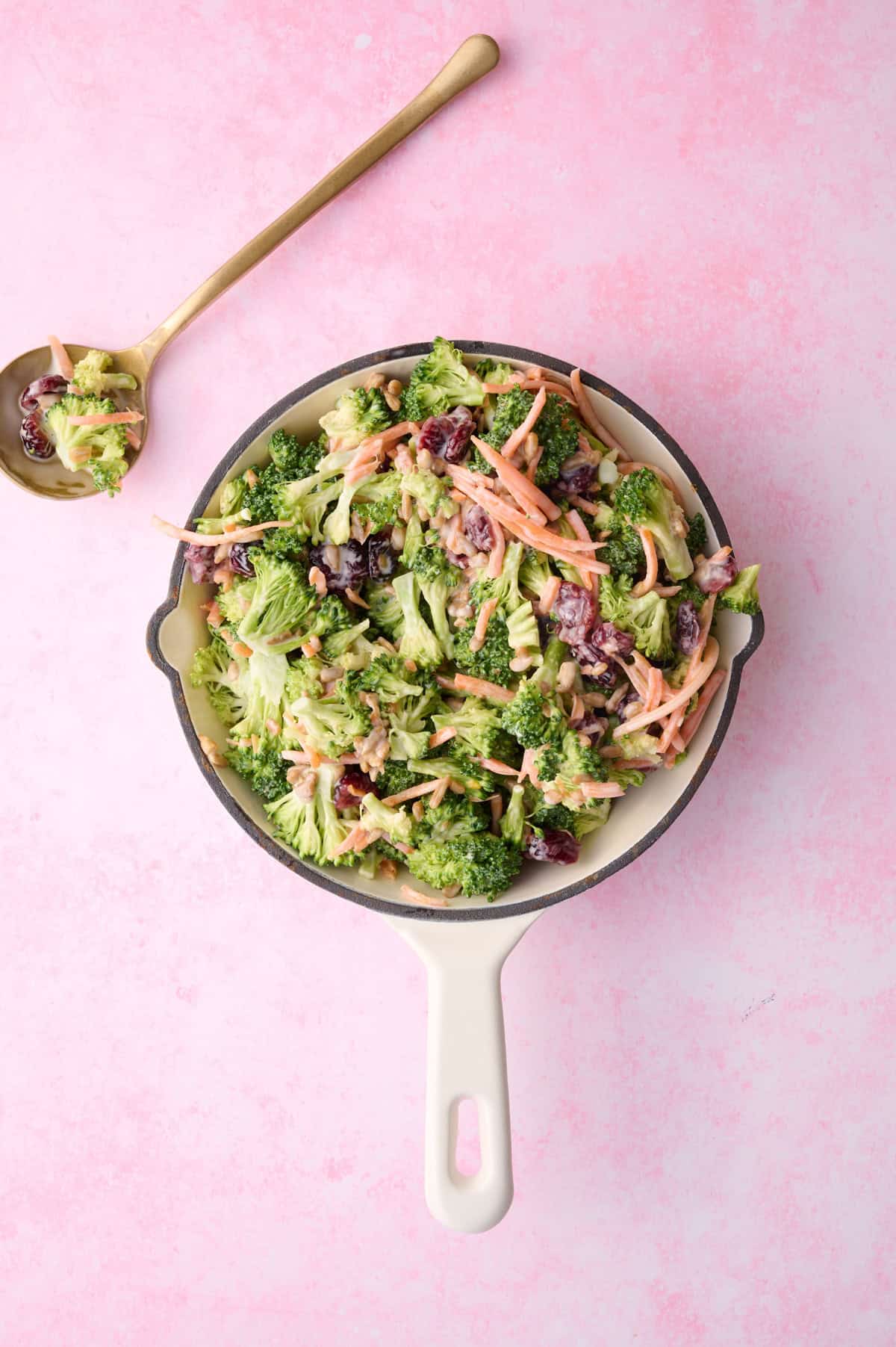 A spoon is holding a little but of the broccoli crunch salad next to a large bowl of the salad.